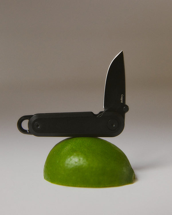 Black pocket knife balanced on a green lime against a neutral background