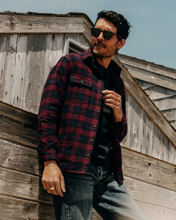 Man wearing a red and black plaid shirt standing in front of a wooden building.